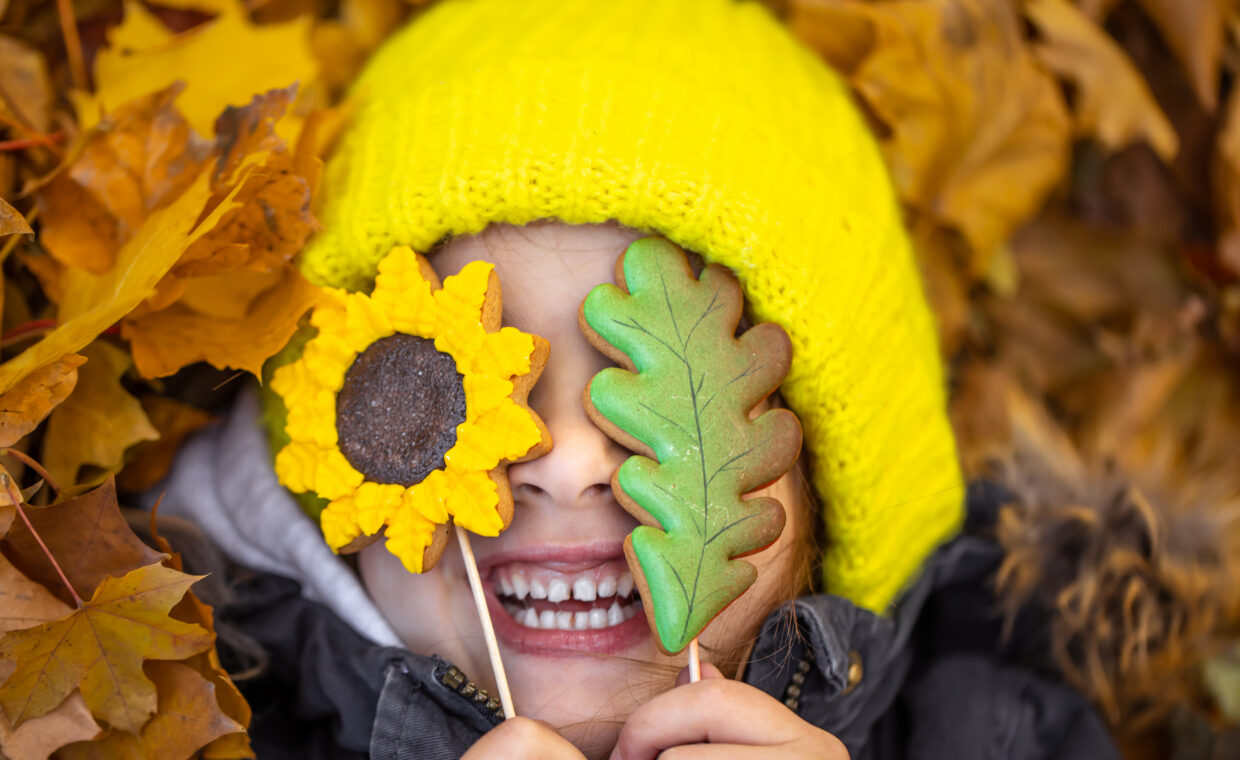 Una bambina con un cappella giallo sdraiata nelle foglie autunnali si copre gli occhi con girasole e foglia verde